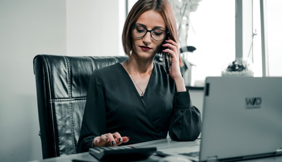 Businesswoman multitasking with a phone and calculator at a desk, showcasing office productivity.
